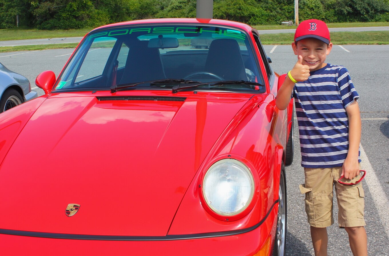 Young boy giving thumbs up beside a red classic sports car.