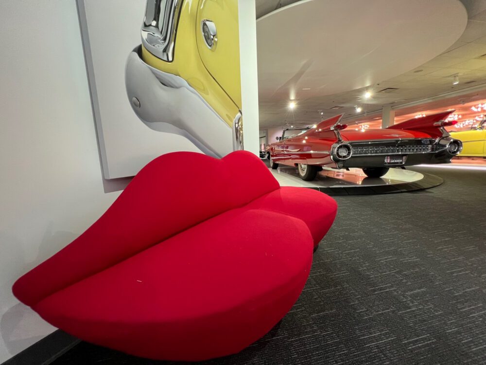 A red lips-shaped sofa in front of classic cars in a museum.