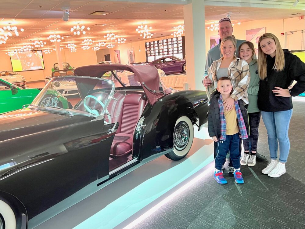 Family posing next to a vintage black convertible car indoors.