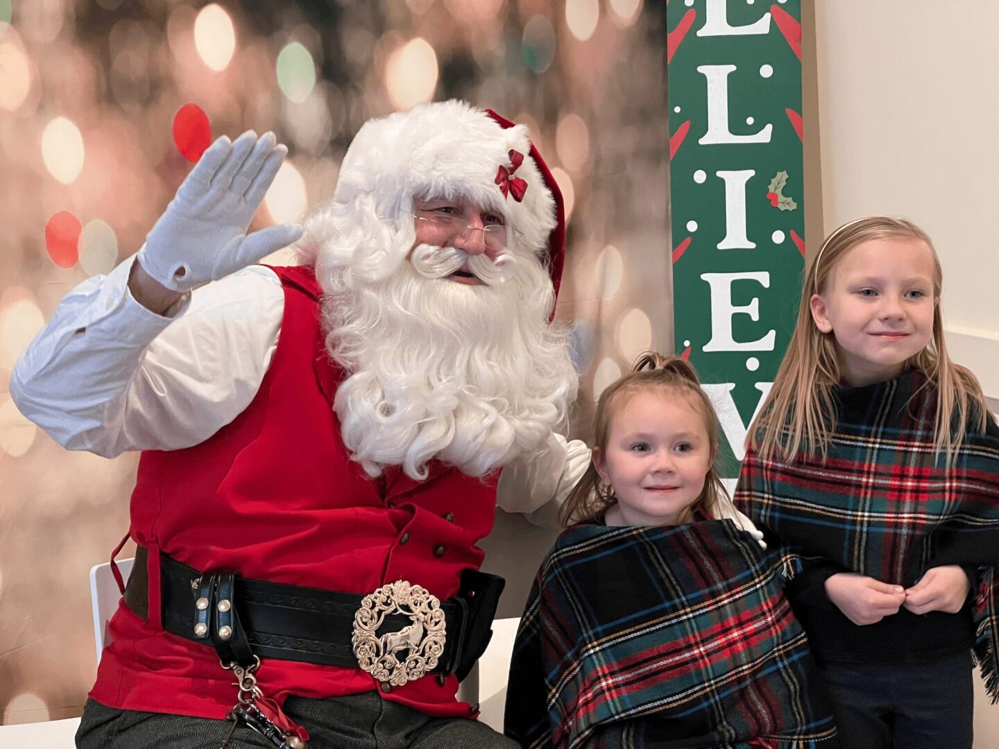 Santa Claus waving with two girls in plaid scarves beside him.