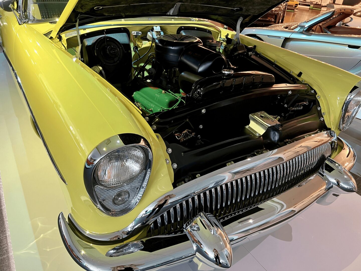 Close-up of a vintage yellow car's front with hood open, revealing the engine.