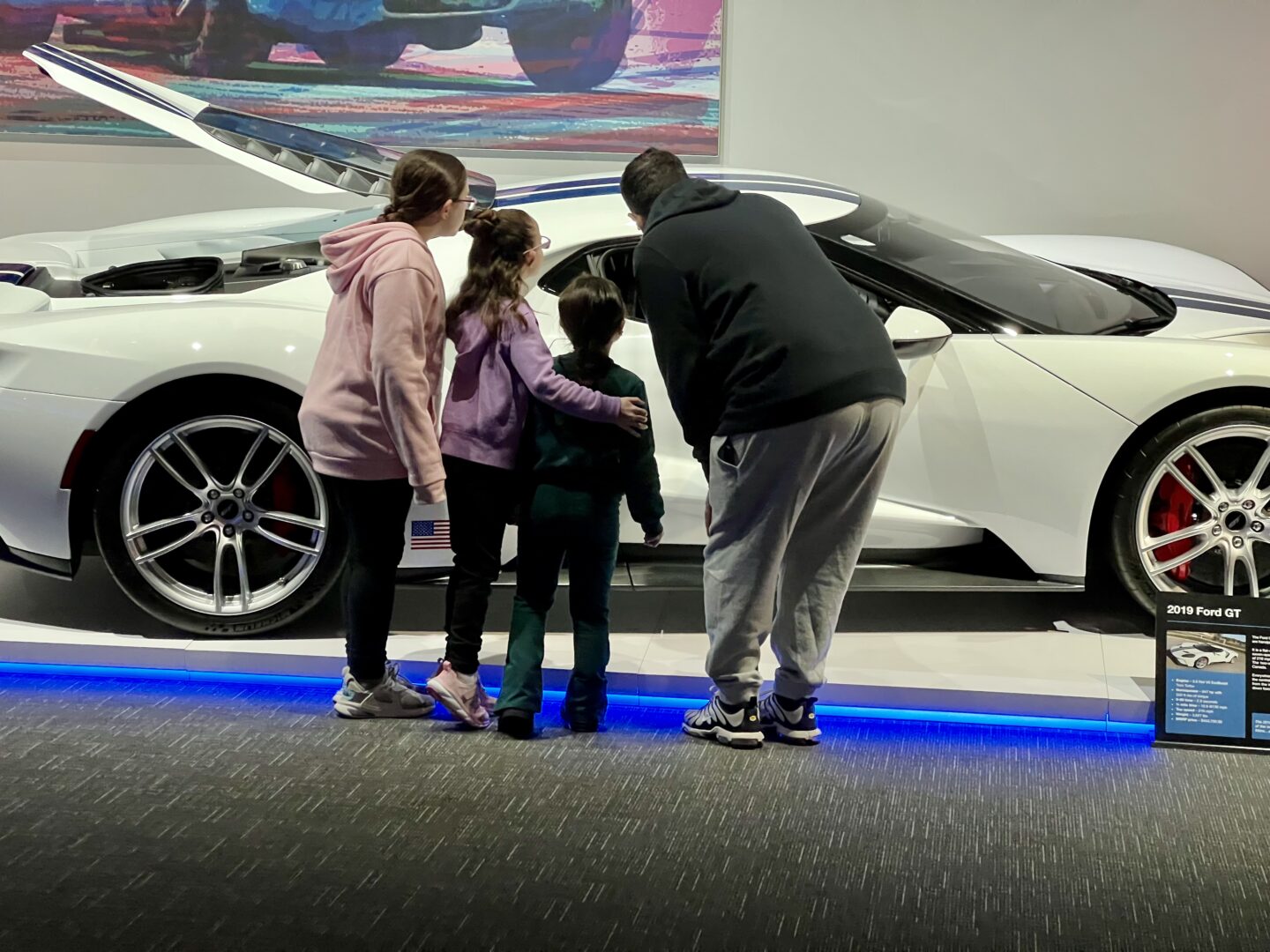 Three children intently observing a white sports car indoors.